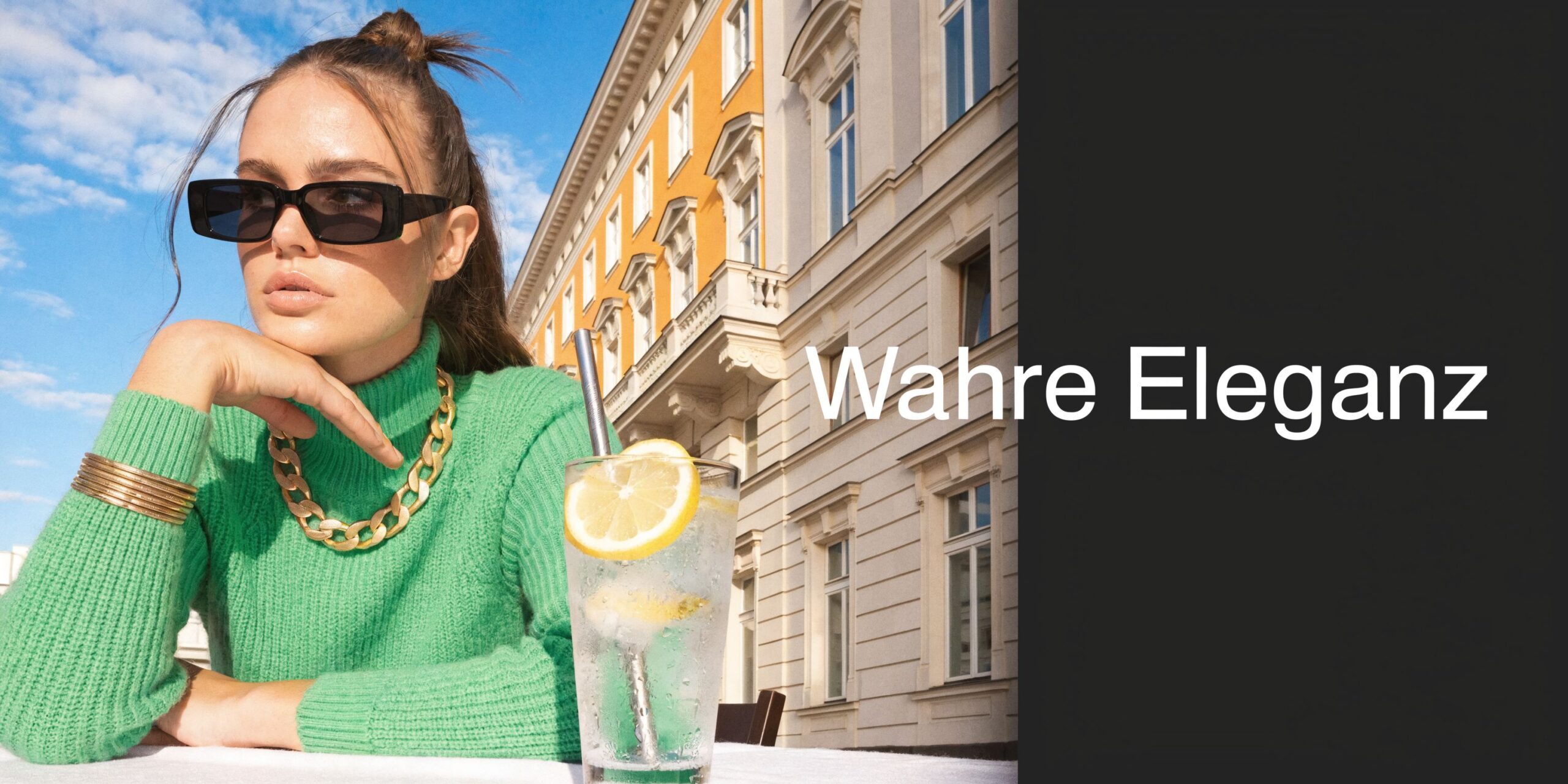 Eine elegante Frau in grünem Pullover sitzt entspannt an einem Tisch vor einem historischen Wiener Gebäude.