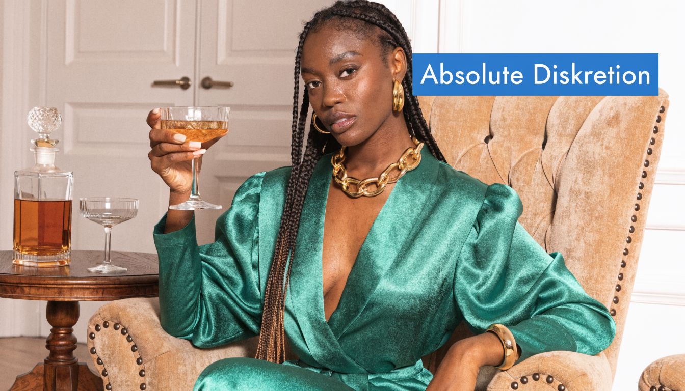 Eine elegante Frau in einem grünen Kleid sitzt mit einem Glas Whisky in einem luxuriösen Sessel.